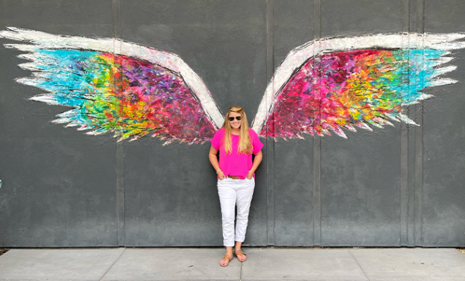 Kasey standing in front of a chalk mural that is a picture of rainbow wings.
