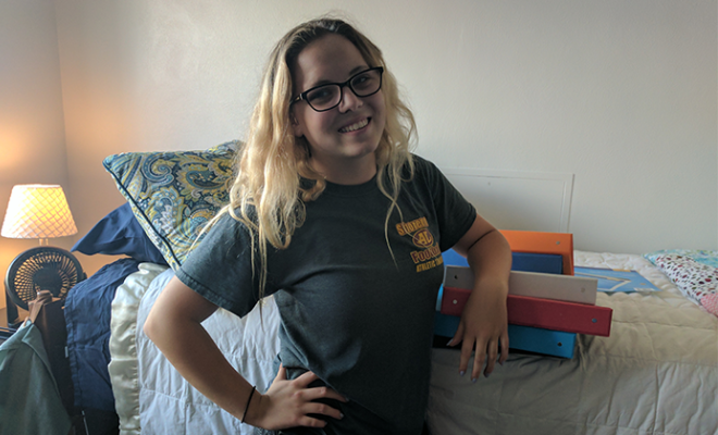 Maddy smiling in her college dorm next to her bed.