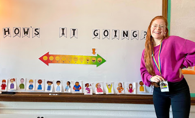 Sarina smiling in front a school white board that says "How's it going?"
